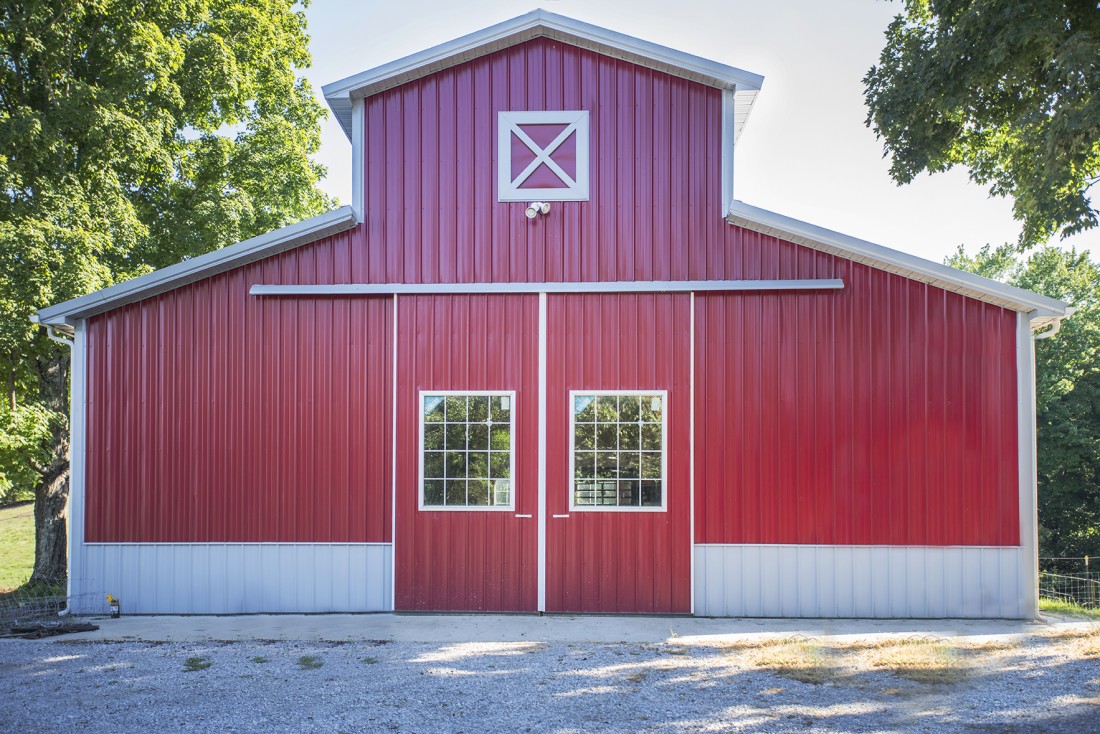 a red barn with metal siding sits in the middle of a tree grove